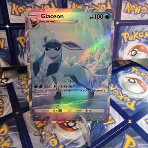 May include: A holographic Glaceon Pok&eacute;mon trading card. The card features a blue and white Glaceon character with the text "Glaceon" and "HP 100". The card has a rainbow holographic effect and is displayed on a clear stand.