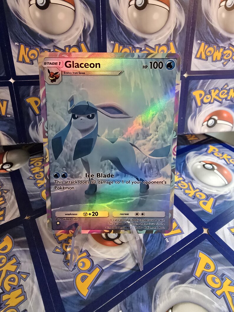 May include: A holographic Glaceon Pok&eacute;mon trading card. The card features a blue and white Glaceon character with the text "Glaceon" and "HP 100". The card has a rainbow holographic effect and is displayed on a clear stand.