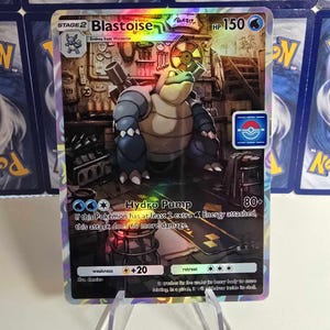 May include: A holographic Blastoise Pok&eacute;mon card. The card features a Blastoise illustration with the text "Hydro Pump" and "HP 150". The card is held in a clear plastic stand, with other Pok&eacute;mon cards visible in the background.