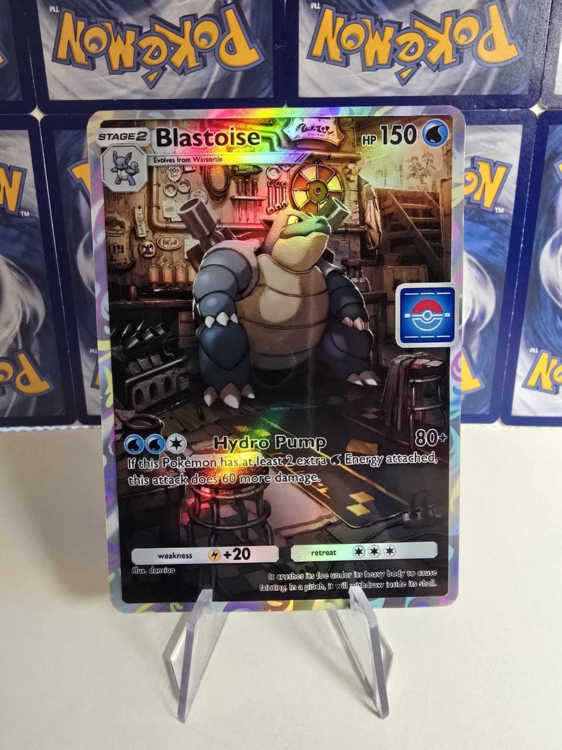 May include: A holographic Blastoise Pok&eacute;mon card. The card features a Blastoise illustration with the text "Hydro Pump" and "HP 150". The card is held in a clear plastic stand, with other Pok&eacute;mon cards visible in the background.
