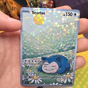 K&ouml;nnte beinhalten: Holografische Snorlax Pok&eacute;mon-Sammelkarte. Die Karte zeigt ein blaues Snorlax, das in einem Feld wei&szlig;er G&auml;nsebl&uuml;mchen unter einer gelben Sonne schl&auml;ft. Der Kartentext enth&auml;lt "Snorlax", "HP 150" und "Collapse".