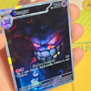 K&ouml;nnte beinhalten: Holografische Gengar Pok&eacute;mon-Karte. Die Karte zeigt ein dunkelviolettes Gengar mit roten Augen und einem breiten Grinsen. Der Text "Gengar" und "Shadow Paralysis" sind vorhanden.