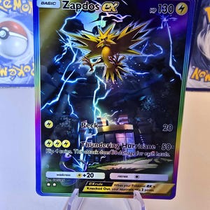 May include: A holographic Zapdos EX Pok&eacute;mon trading card. The card features a yellow bird-like creature with lightning effects. Text includes "Zapdos EX", "HP 130", and attack descriptions. The card is displayed in a clear plastic holder.