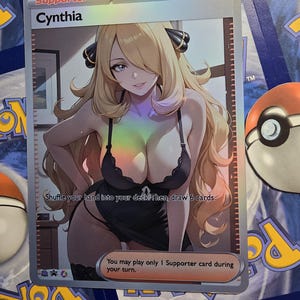 Op de afbeelding: Een holografische Cynthia Trainerkaart uit het Pok&eacute;mon Trading Card Game. De kaart toont een illustratie van een vrouw met blond haar in een zwarte outfit. De kaarttekst bevat "Schud je hand in je deck. Trek dan 6 kaarten."