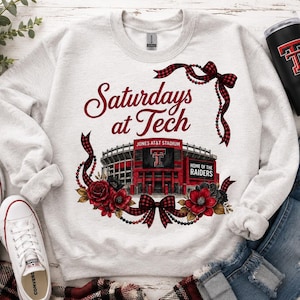 May include: A light gray crewneck sweatshirt featuring a graphic of Jones AT&T Stadium with the text "Saturdays at Tech" and "Home of the Raiders," accented by red and black buffalo plaid ribbons, beads, and floral illustrations in red and black.