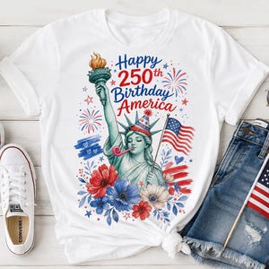 Peut inclure: Un t-shirt blanc &agrave; manches courtes orn&eacute; d'une illustration de la Statue de la Libert&eacute; portant un chapeau de f&ecirc;te et soufflant dans une sarbacane. Le texte indique Happy 250th Birthday America, avec des feux d'artifice, des fleurs et un drapeau.