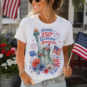 Peut inclure: Un t-shirt blanc &agrave; manches courtes avec un motif repr&eacute;sentant la Statue de la Libert&eacute; portant un chapeau de f&ecirc;te et soufflant dans une sarbacane. Le design inclut des fleurs rouges, blanches et bleues, des feux d'artifice et le texte Happy 250th Birthday America.
