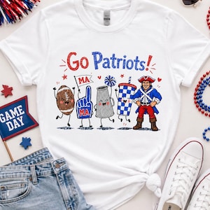 May include: A white short-sleeved t-shirt featuring a graphic print with the text "Go Patriots!" and cartoon characters including a football, a foam finger, a megaphone, a checkered cup, and a mascot dressed in a colonial-style uniform.