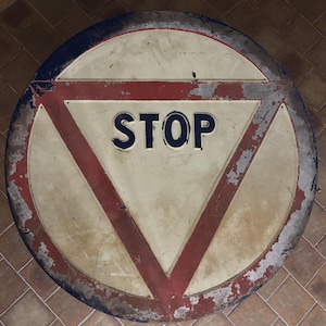 Puede incluir: Un letrero de stop redondo y desgastado, con un centro blanco y un borde rojo. La palabra "STOP" está en letras negras en negrita. El letrero tiene una apariencia vintage y desgastada con pintura desconchada.