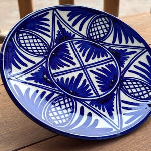 Peut inclure: Une assiette ronde en céramique avec un motif floral bleu et blanc. L'assiette présente un motif central en forme d'éclat d'étoile avec des éléments floraux et géométriques rayonnants. Le bord est bleu foncé, et l'assiette est posée sur une surface en bois.