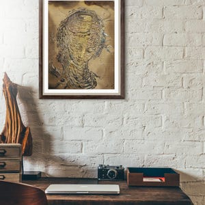 K&ouml;nnte beinhalten: Ein gerahmtes Kunstwerk, das ein stilisiertes Gesicht mit komplizierten, geschichteten Details in Gold- und Braunt&ouml;nen zeigt. Das Kunstwerk wird an einer wei&szlig;en Backsteinmauer &uuml;ber einem Holzschreibtisch mit Laptop, Kamera und Holzaccessoires ausgestellt.