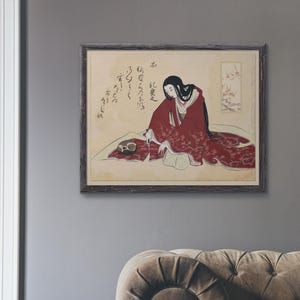 Könnte beinhalten: Gerahmter Druck eines japanischen Kunstwerks. Eine Frau in einem roten Kimono mit floralen Mustern sitzt auf einer Matte. Das Kunstwerk zeigt japanische Kalligraphie und ein kleines Blumendesign. Der Rahmen ist dunkelbraun.
