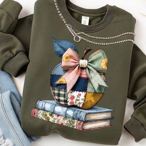 Könnte beinhalten: Olivgrünes Sweatshirt mit einem Apfeldesign aus Patchworkstoff, einer großen Schleife und einer Kettenverzierung. Der Apfel liegt auf einem Stapel Bücher. Hellblaue Jeans sind zu sehen. Ein lässiger, bequemer Stil.
