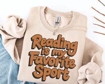 May include: A beige sweatshirt with the text "Reading is my Favorite Sport" in brown and black lettering. The sweatshirt is folded, with a silver necklace draped across it. A pair of blue jeans and a beige sweater are visible in the background.