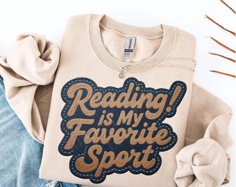 May include: A beige sweatshirt with the text "Reading is My Favorite Sport" in a stitched-on design. The text is in brown and navy blue, with a light blue stitch outline. A silver necklace is draped on the sweatshirt.