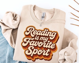 May include: A beige sweatshirt with the text "Reading is my Favorite Sport" in a retro font. The text is in shades of brown and orange. A silver necklace is draped on the sweatshirt.
