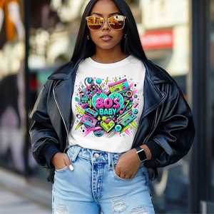May include: White t-shirt with a colorful 80's-themed graphic. The design features a heart shape with the text "80's BABY" and images of cassette tapes, boomboxes, and geometric shapes in neon colors. The person is wearing a black leather jacket and blue jeans.