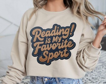 May include: Beige sweatshirt with the text "Reading is My Favorite Sport" in a brown and blue design. The sweatshirt is a neutral color and has a crew neck.