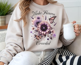 Könnte beinhalten: Beiges Sweatshirt mit einem Blumendesign mit lila Sonnenblumen und Schmetterlingen. Der Text "Make Heaven Crowded" ist oberhalb und unterhalb des Blumenarrangements aufgedruckt. Die Person hält einen weißen Becher.