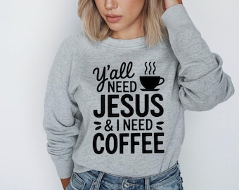 Może przedstawiać: Jasnoszara bluza z napisem "Y'all NEED JESUS & I NEED COFFEE" w kolorze czarnym. Projekt zawiera grafikę filiżanki kawy. Bluza jest w stylu casual.