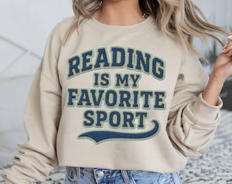 May include: Beige crewneck sweatshirt with the text "READING IS MY FAVORITE SPORT" in blue and light blue lettering. The sweatshirt has a relaxed fit and is made of a soft material.