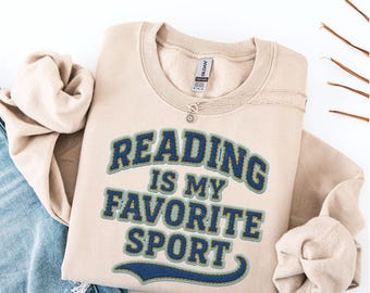 May include: A beige sweatshirt with the text "READING IS MY FAVORITE SPORT" in blue and gold lettering. The sweatshirt is folded on a white surface, with a silver necklace and a pair of jeans visible. The sweatshirt is a casual style.