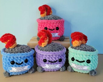 May include: A collection of handmade, colorful crochet "Emotional Support Dumpster Fire" plush toys. Each plush is a different color, including blue, pink, purple, and green, with a gray top and a red and orange flame. They have smiling faces and small tags.