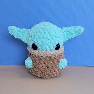 May include: A plush, light blue and brown crocheted character with large ears and black eyes. The character is a popular science fiction figure, and the plush is made of soft, textured yarn. The character's head and ears are light blue, and the body is brown.
