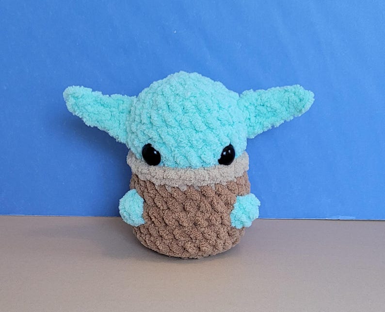 May include: A plush, light blue and brown crocheted character with large ears and black eyes. The character is a popular science fiction figure, and the plush is made of soft, textured yarn. The character's head and ears are light blue, and the body is brown.