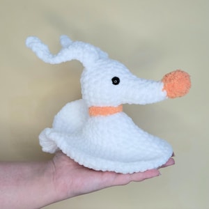 May include: A white plush toy of a ghost dog with a long snout and floppy ears. The dog has a black eye and an orange nose and collar. The toy is held in a hand against a light yellow background.