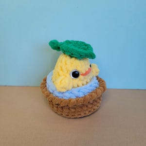 May include: A handmade, plush, yellow pear-shaped creature with large black eyes, a small orange mouth, and a green leaf on its head. It sits in a brown basket with a blue lining, all made of soft yarn.