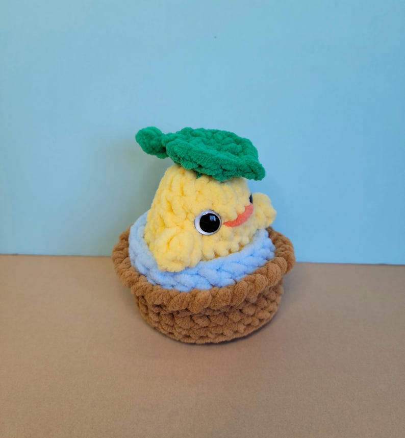 May include: A handmade, plush, yellow pear-shaped creature with large black eyes, a small orange mouth, and a green leaf on its head. It sits in a brown basket with a blue lining, all made of soft yarn.