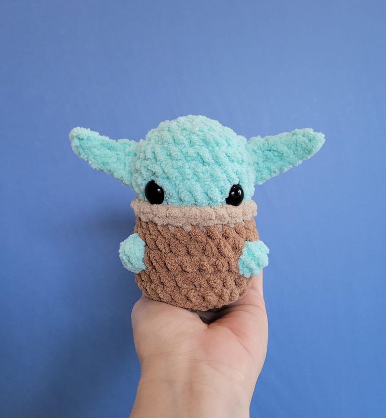 May include: A handmade plush doll in the likeness of a character from a popular science fiction series. The doll features a light blue head and ears, black eyes, and a brown and tan body. The doll is made of a soft, textured material.
