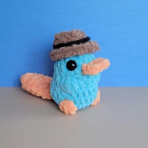 May include: A handmade, plush platypus toy in shades of blue, peach, and brown. The toy has a light blue body, peach-colored feet, bill, and tail, and a brown hat with a black band. The eyes are black.