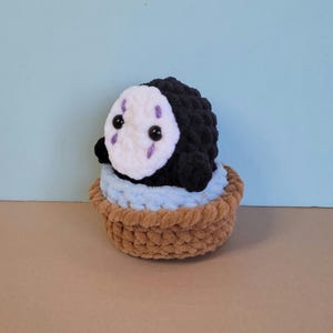 May include: A handmade crochet plushie of a character from Spirited Away. The black plushie has a white face with purple markings and black eyes. It sits in a brown basket with a light blue lining, against a light blue background.
