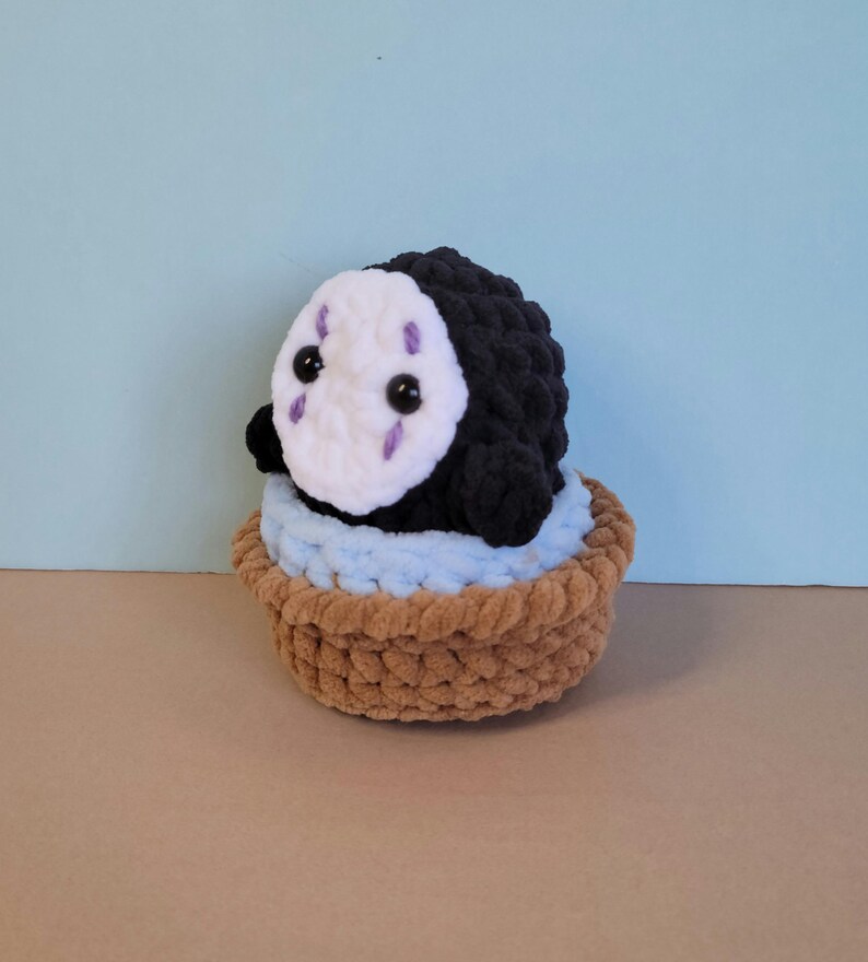 May include: A handmade crochet plushie of a character from Spirited Away. The black plushie has a white face with purple markings and black eyes. It sits in a brown basket with a light blue lining, against a light blue background.