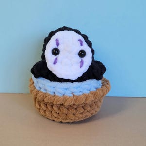 May include: A handmade crochet plush toy of the character No-Face from Spirited Away. The plush features a white face with black eyes and purple markings, a black body, and sits in a brown and blue basket. The toy is made of soft yarn.