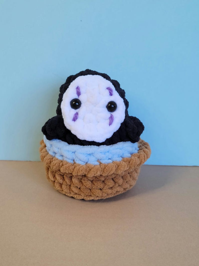 May include: A handmade crochet plush toy of the character No-Face from Spirited Away. The plush features a white face with black eyes and purple markings, a black body, and sits in a brown and blue basket. The toy is made of soft yarn.