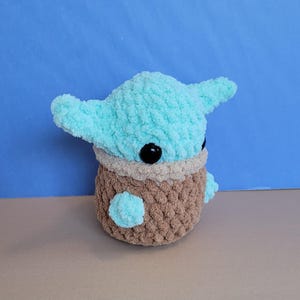 May include: A handmade plush toy featuring a character with large ears and black eyes. The head is light blue, the body is brown, and the eyes are black. The toy is made of a soft, textured yarn.