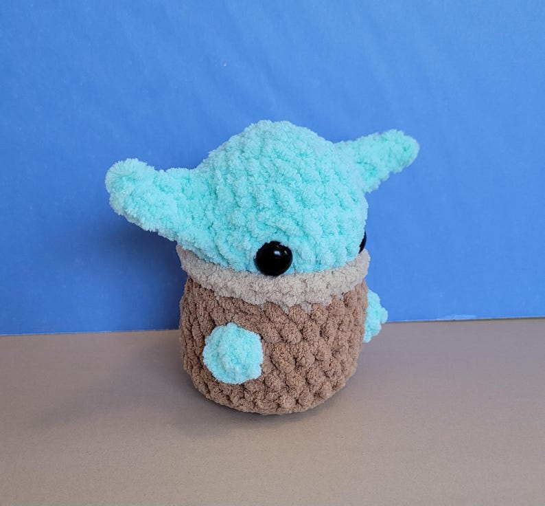 May include: A handmade plush toy featuring a character with large ears and black eyes. The head is light blue, the body is brown, and the eyes are black. The toy is made of a soft, textured yarn.