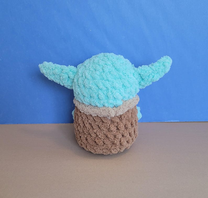May include: A handmade plush toy in the likeness of a character with large ears. The toy features a light blue head and ears, a tan neck, and a brown body. The plush is made of a soft, textured yarn.