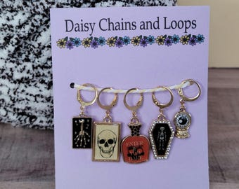 May include: A set of five gold-toned metal stitch markers on a light purple display card labeled Daisy Chains and Loops. The charms include a skeletal hand, a skull, a potion bottle with the text Enter If You Dare, a skeleton in a coffin, and an eye.