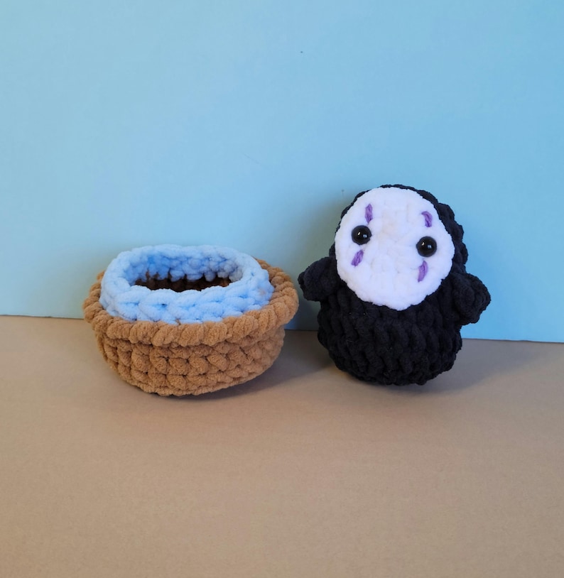 May include: Crocheted basket with a light blue interior and a brown exterior, next to a black and white crocheted character with a mask-like face and purple accents. The items are handmade.