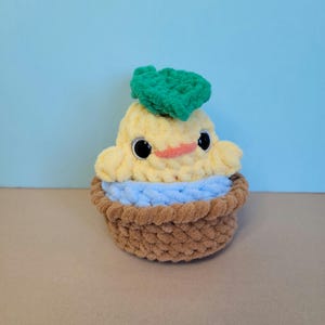 May include: A handmade, plush, yellow and blue cupcake-shaped toy with a smiling face and a green leaf on top. The toy has large black eyes and a small orange beak. The cupcake sits in a brown basket.