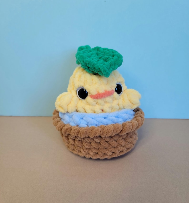 May include: A handmade, plush, yellow and blue cupcake-shaped toy with a smiling face and a green leaf on top. The toy has large black eyes and a small orange beak. The cupcake sits in a brown basket.