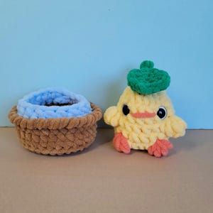 May include: A handmade, yellow crocheted duck with a green beret and a small brown basket with a blue interior. The duck has black eyes and orange feet and beak. The basket is brown.