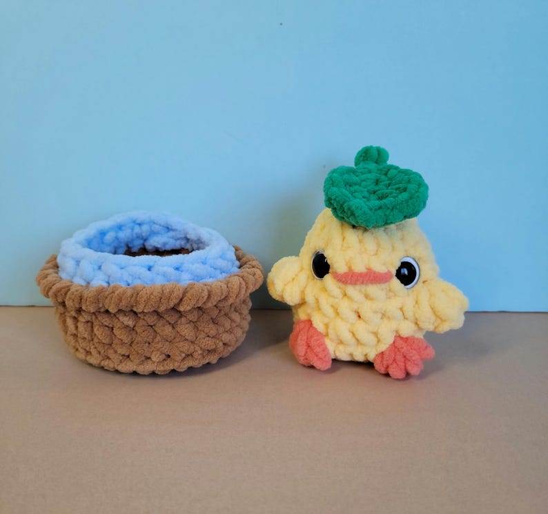 May include: A handmade, yellow crocheted duck with a green beret and a small brown basket with a blue interior. The duck has black eyes and orange feet and beak. The basket is brown.
