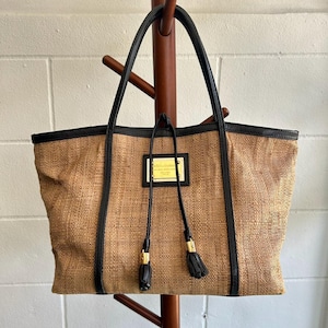 May include: A brown and black woven tote bag with black leather trim and gold accents. The bag has a large, rectangular shape and two long handles. The bag has a gold label with the text "Dolce & Gabbana" on the front.