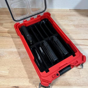 May include: Open red and black tool organizer with a clear lid. The lid has the Milwaukee logo and text. The interior has several black compartments holding black tools. The organizer has a black handle and metal latches.
