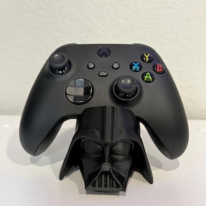 May include: Black Xbox controller resting on a black Darth Vader-themed stand. The controller has colored buttons and a directional pad. The stand features a detailed depiction of Darth Vader's helmet.